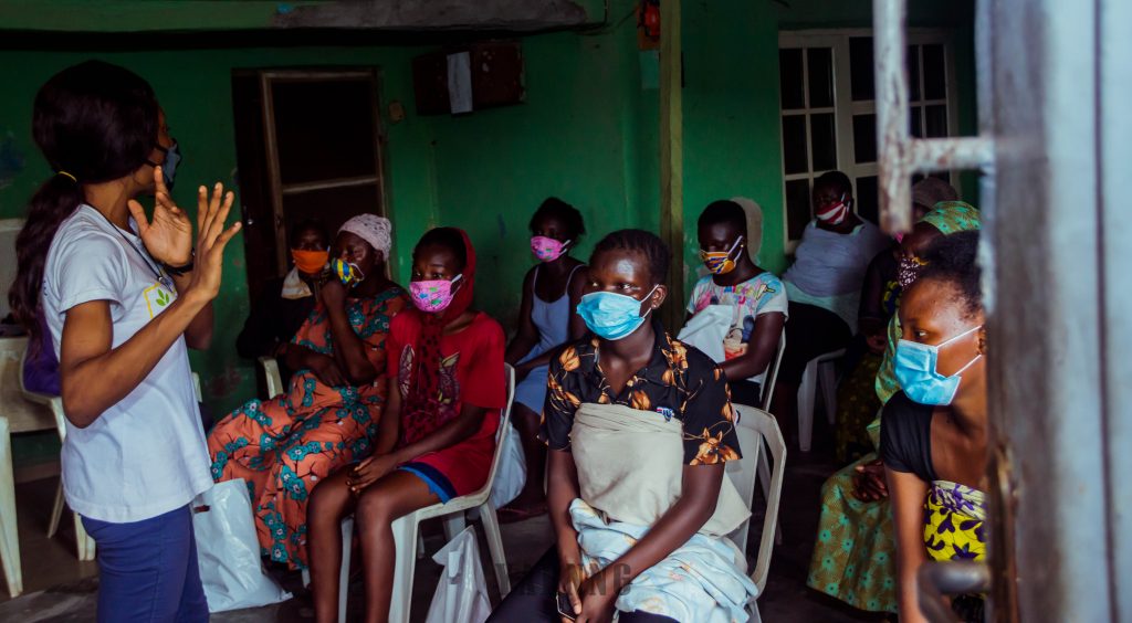 Image of a young female addressing a group of girls and women sitting on white chairs wearing face masks while the person talking to them is demonstrating with her hands.